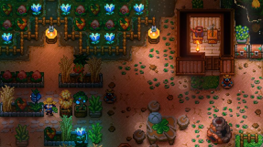 A top-down farming simulation gameplay screenshot from Core Keeper, showing an underground base setup with crop plots growing blue flowers and other vegetation, wooden fencing, lighting elements, a small cabin with crafting stations, and a character exploring the darkened cavern surroundings.