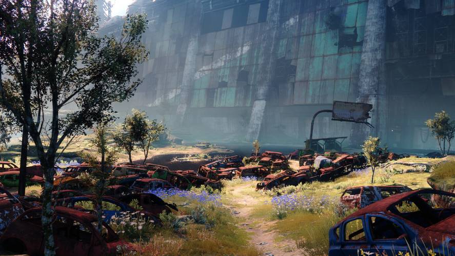 A post-apocalyptic gameplay screenshot from Destiny 2, showing a grassy, overgrown path winding through a field of rusted, abandoned cars and wildflowers, with towering, weathered concrete and metal ruins looming in the background under a bright sky.