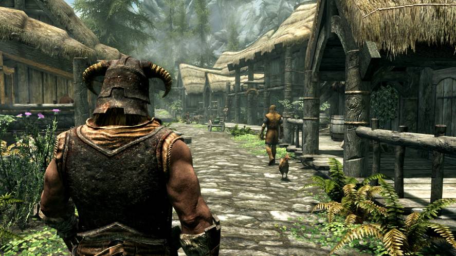 A third-person screenshot from the video game Skyrim, showing the back of a character wearing an Iron Helmet and armor, walking down a stone path through a village with thatched-roof wooden buildings, lush green ferns, and a villager walking ahead with a chicken.